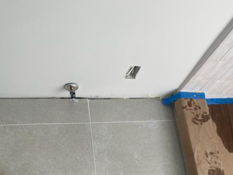 A water shutoff valve stubbed out at the base of a white wall, above a new gray tile floor partly covered with protective paper