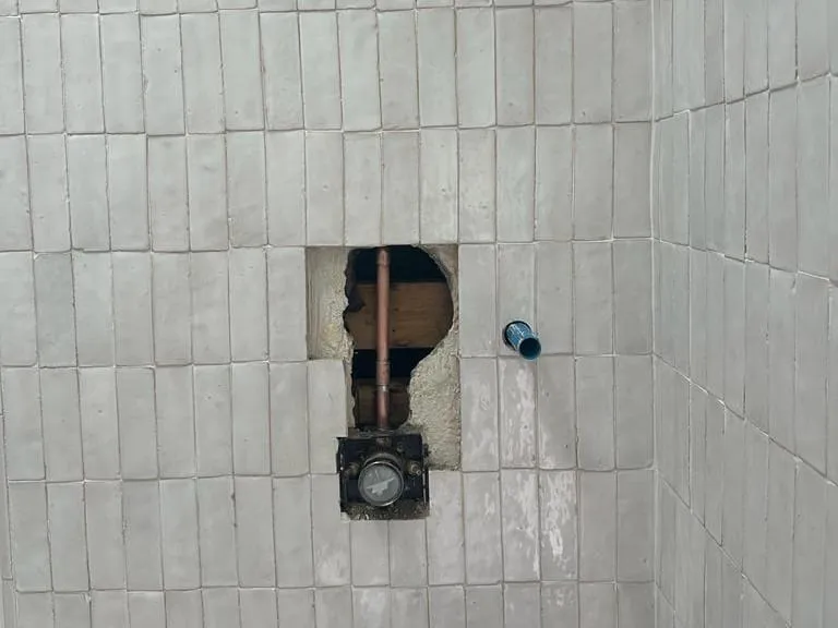 An opening cut in a white tiled shower wall to reach the valve and copper pipe behind it